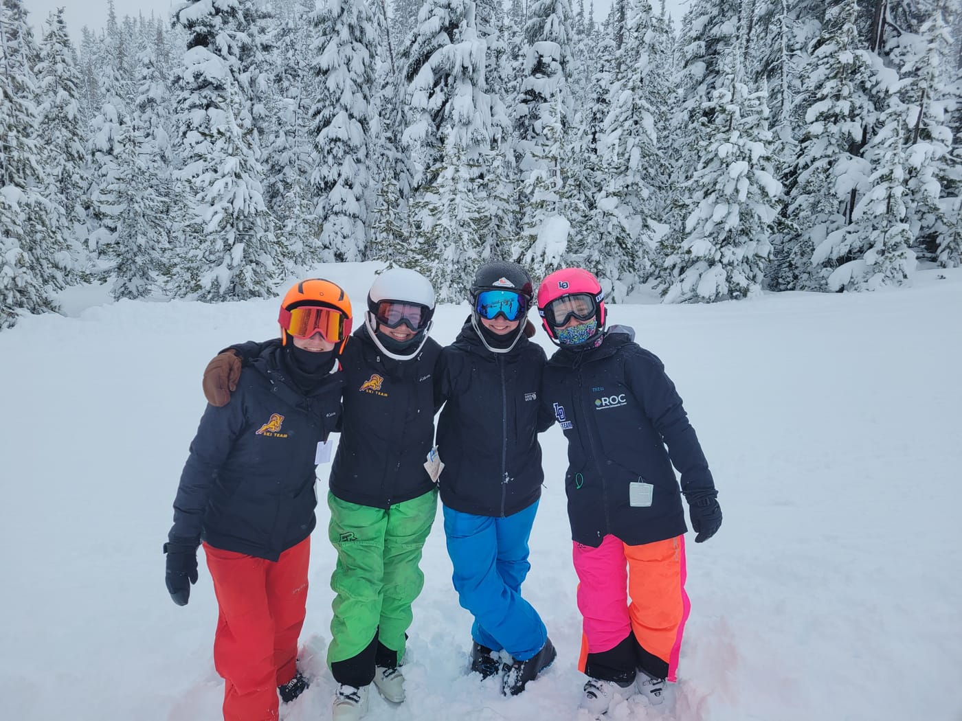 Four racers arm in arm in front of snow-loaded firs, ski pants in orange, green, blue and pink